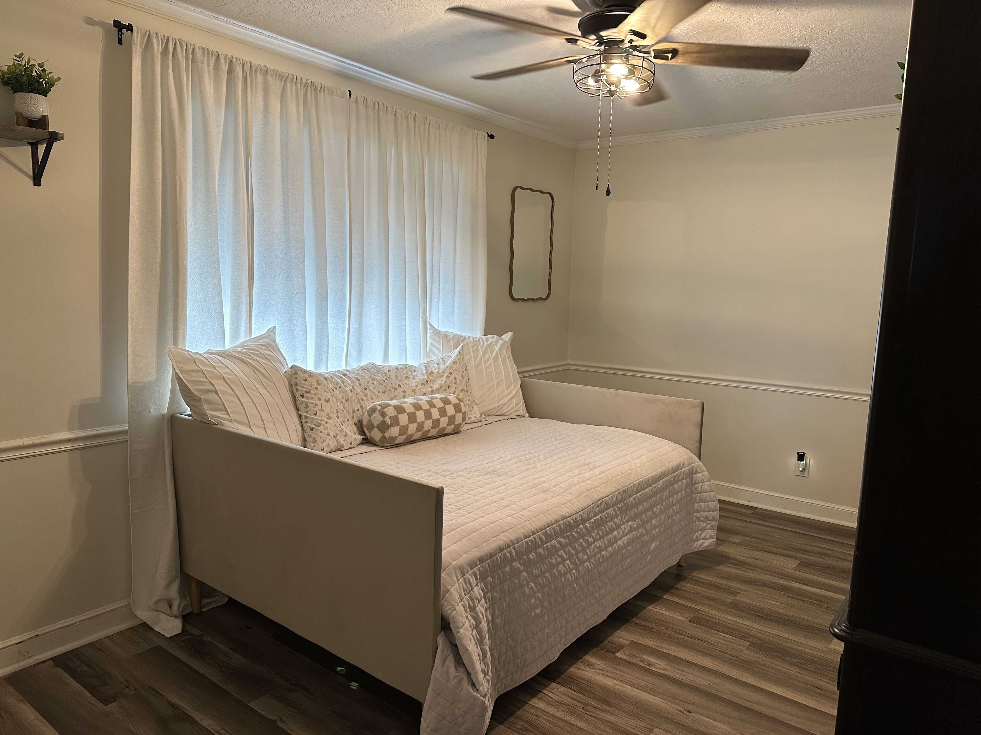 Ein Schlafzimmer mit Bett, Deckenventilator, Fenster mit Vorhängen und einem wandmontierten Spiegel.
