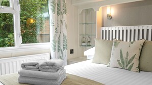 A neatly made bed with a folded towel, a bedside table with a glass shelf, and a window with curtains. - Firgarth Lodge  - sleeps 6 guests  in 3 bedrooms (Windermere)