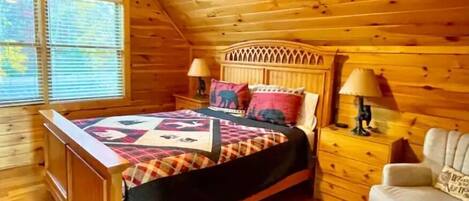 Quarto de cabana de madeira aconchegante, com uma cama, duas mesas de cabeceira, uma cadeira e uma janela com persianas.
