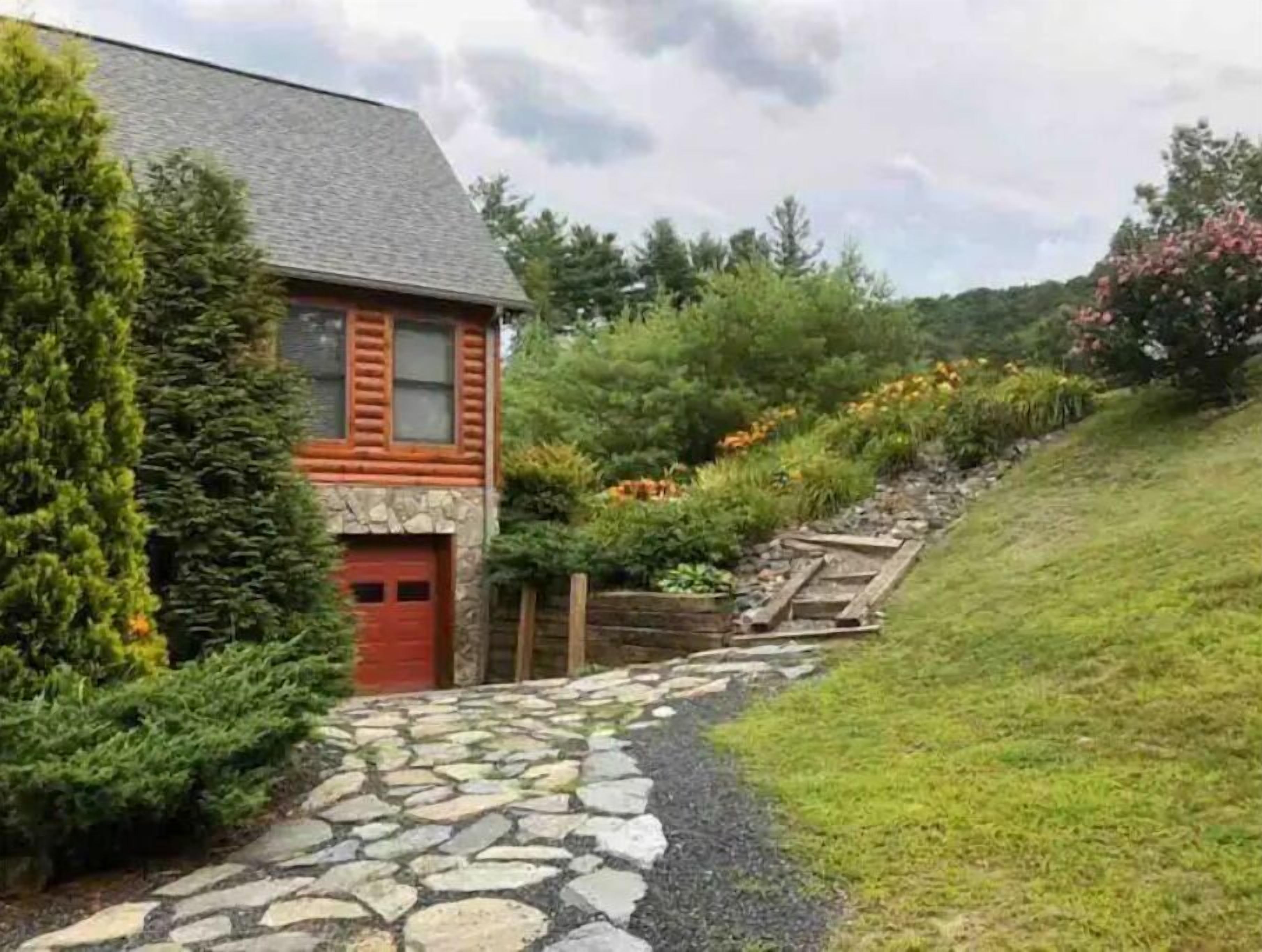 A log cabin with a stone pathway leading to the entrance, surrounded by lush greenery and flowering plants.