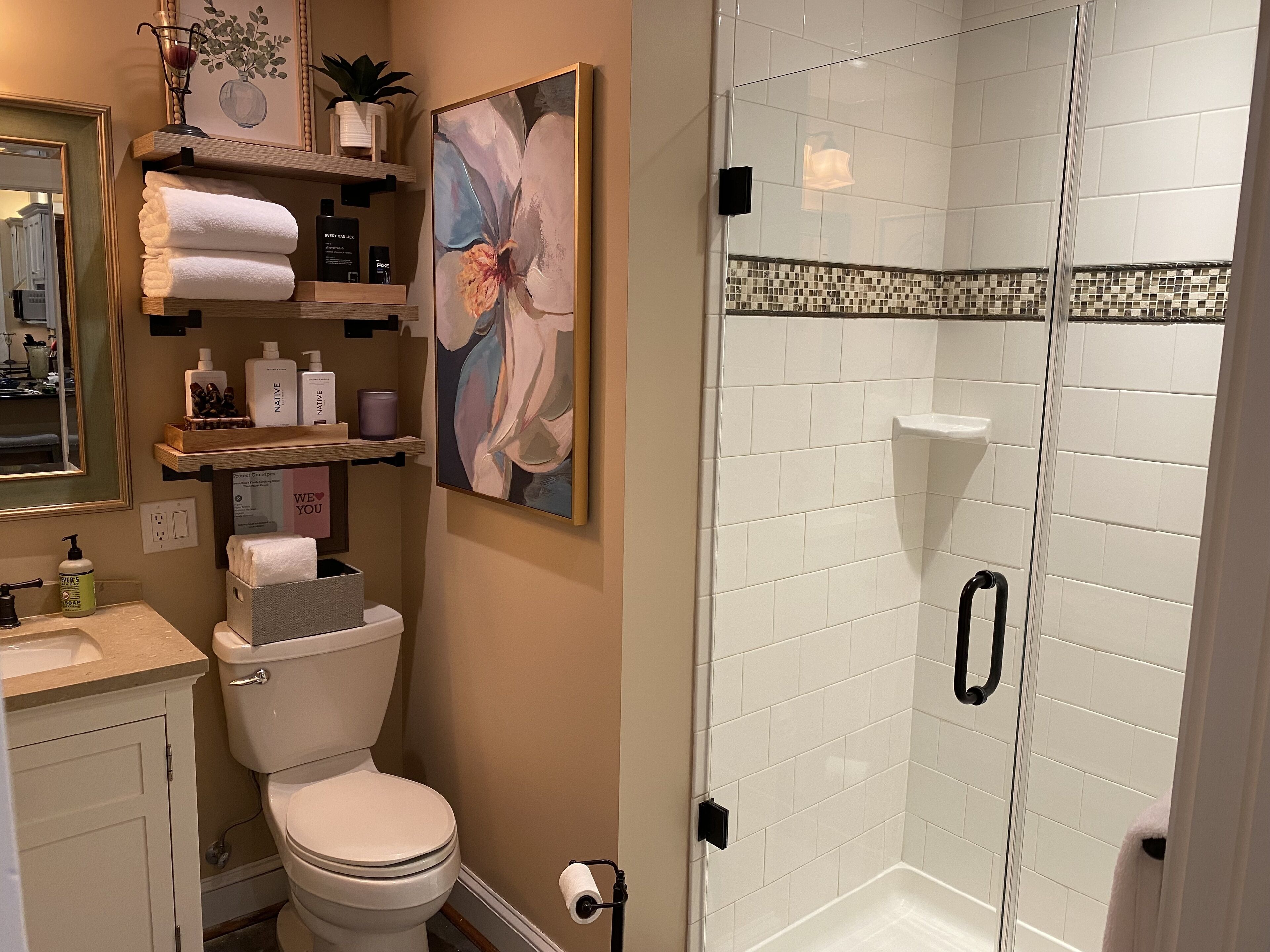 Ein Badezimmer mit Toilette, Waschbecken und Dusche. Es gibt Regale mit Toilettenartikeln und ein gerahmtes Bild an der Wand.