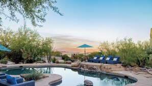 A pool area with lounge chairs and umbrellas, surrounded by desert vegetation and a view of distant mountains.
