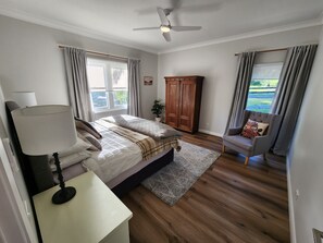 A bedroom with a bed, a bedside table with a lamp, a wooden wardrobe, a chair, and a view of the outdoors.