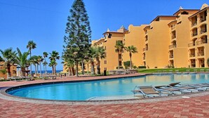 Ein Poolbereich eines Resorts mit klarem, blauem Pool, Palmen und einem großen Gebäude mit Balkonen.