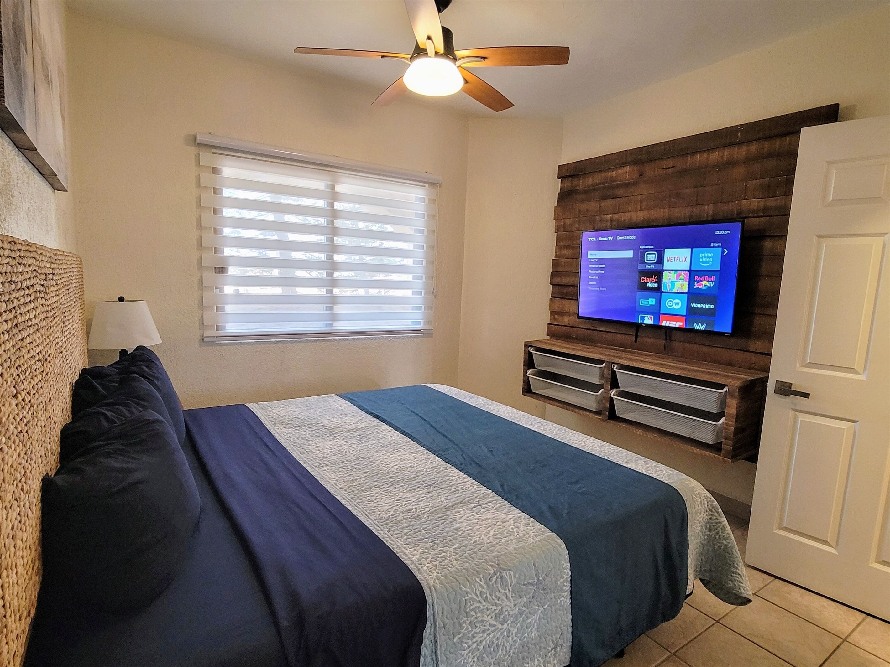 Une chambre à coucher comprenant un lit, un ventilateur de plafond, un téléviseur fixé au mur, une fenêtre avec des stores et une porte.