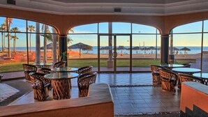 A spacious indoor area with large windows offering a view of the beach and ocean. There are several round tables and wicker chairs arranged for dining.