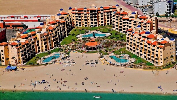 An aerial view of a resort with multiple buildings, a large sandy beach, and several swimming pools.