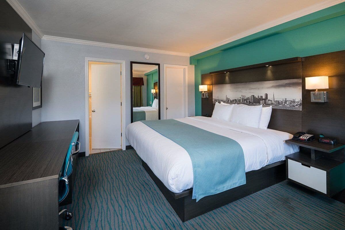 Une chambre d’hôtel comprenant un lit, une table de chevet, un bureau, une chaise et un tableau représentant un paysage urbain accroché au mur.
