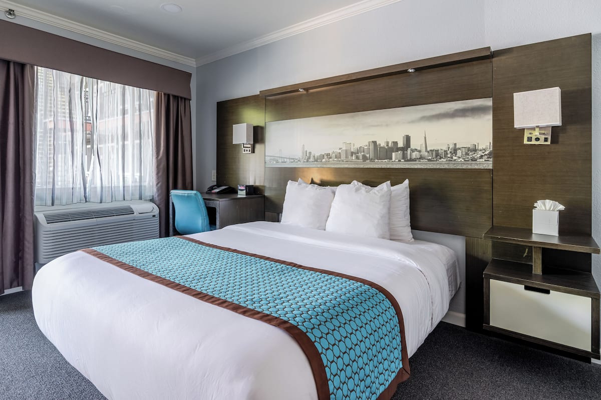 Une chambre d’hôtel avec un grand lit, un tableau représentant un paysage urbain, un bureau avec un téléphone et une table de chevet avec une boîte de mouchoirs.