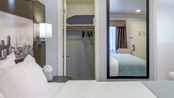 Quarto de hotel com cama, mesa de cabeceira, abajur, cabideiro e porta que dá para outro cômodo.