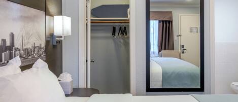 A hotel room with a bed, a nightstand, a lamp, a coat rack, and a door leading to another room.