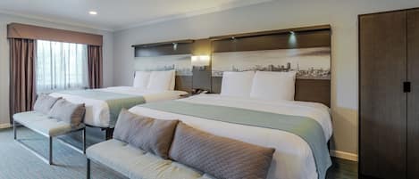Une chambre d’hôtel avec deux lits, un banc, une œuvre d’art représentant un paysage urbain au niveau de la tête de lit et une armoire.