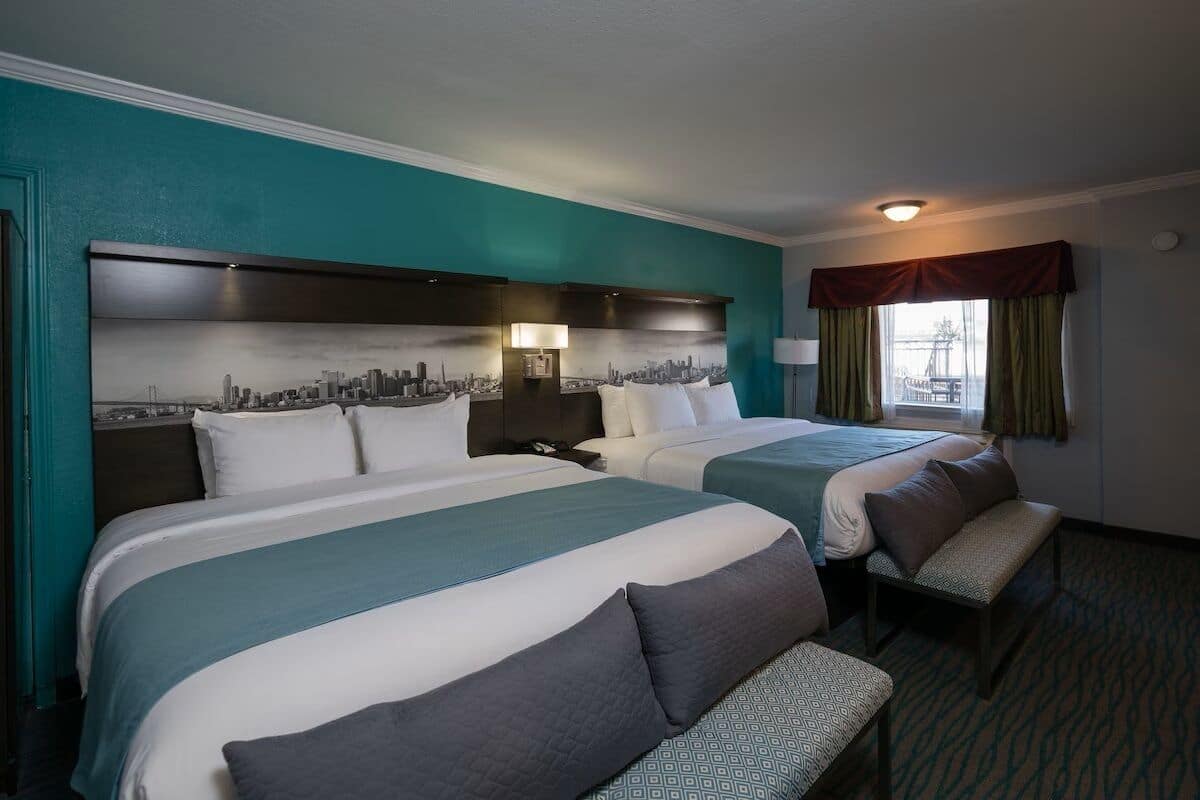 Une chambre d’hôtel avec deux lits, des murs de couleur turquoise, une fresque représentant un paysage urbain sur la tête de lit, un banc et une fenêtre avec des rideaux.