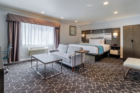 Una habitación de hotel con cama, sofá, una mesita, una silla y ventana con cortinas.. Symphony Inn