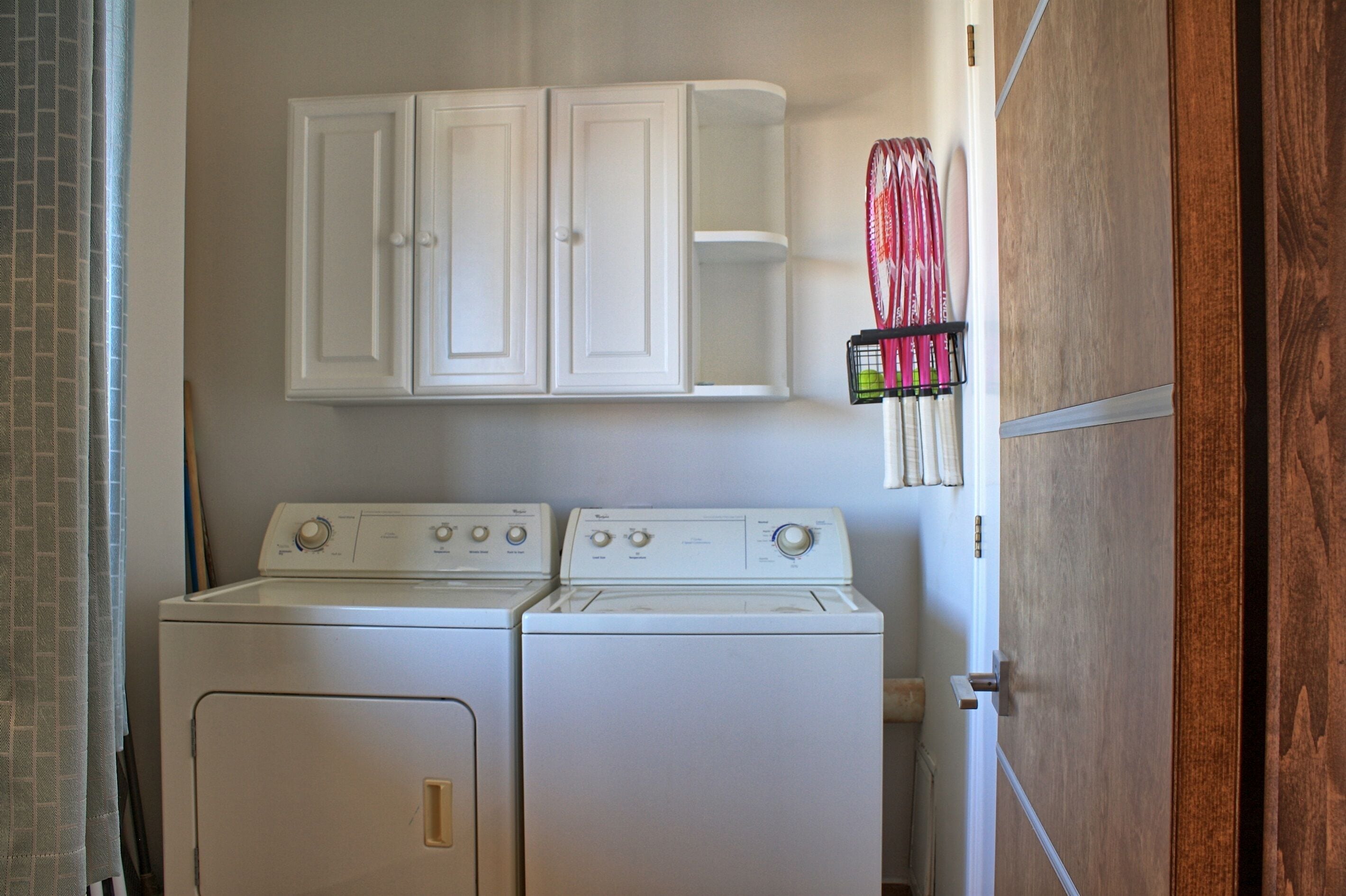 Un espace buanderie comprenant deux lave-linge à chargement frontal blancs, un meuble blanc au-dessus et un porte-raquettes fixé au mur.