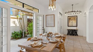 A dining area with a long table set for six, surrounded by chairs, with a chandelier above and a framed artwork on the wall.