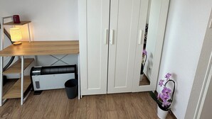 Una habitación pequeña y bien iluminada con un armario blanco, un escritorio con lámpara, un calefactor y una orquídea en maceta.