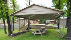 Et overdækket picnicområde med et bord og bænke, omgivet af træer og en bygning i baggrunden.