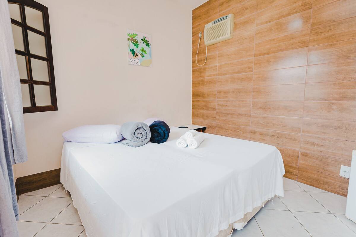 A hotel room with a bed, a towel, a pillow, a wall-mounted air conditioner, and a small artwork.