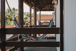 Un balcón de madera con hamacas y vistas a palmeras y un edificio con techo de tejas.