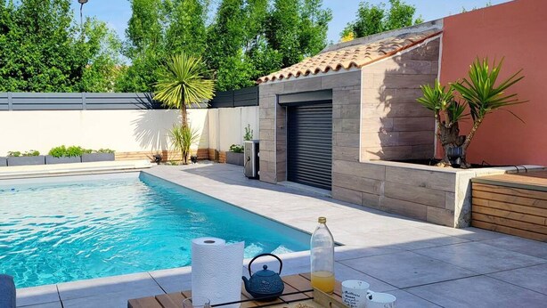 A poolside area with a wooden table set for breakfast, a bottle of juice, and a teapot. - Villa proche de La Rochelle avec piscine (VILLEDOUX)
