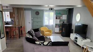 A living room with a curved sofa, a yellow armchair, a flat-screen TV, a bookshelf, and a dining area with a table and chairs. - Villa proche de La Rochelle avec piscine (VILLEDOUX)