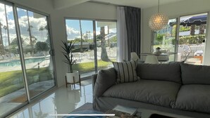 A modern living room with large windows, a gray sofa, and a view of a pool and outdoor seating area. - Iconic Mid-Century Modern Gem – Featured on Palm Springs Modernism Week Tour! (Palm Springs)