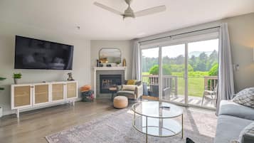 A modern living room with a flat-screen TV, a fireplace, a balcony with a view of greenery, and a ceiling fan.