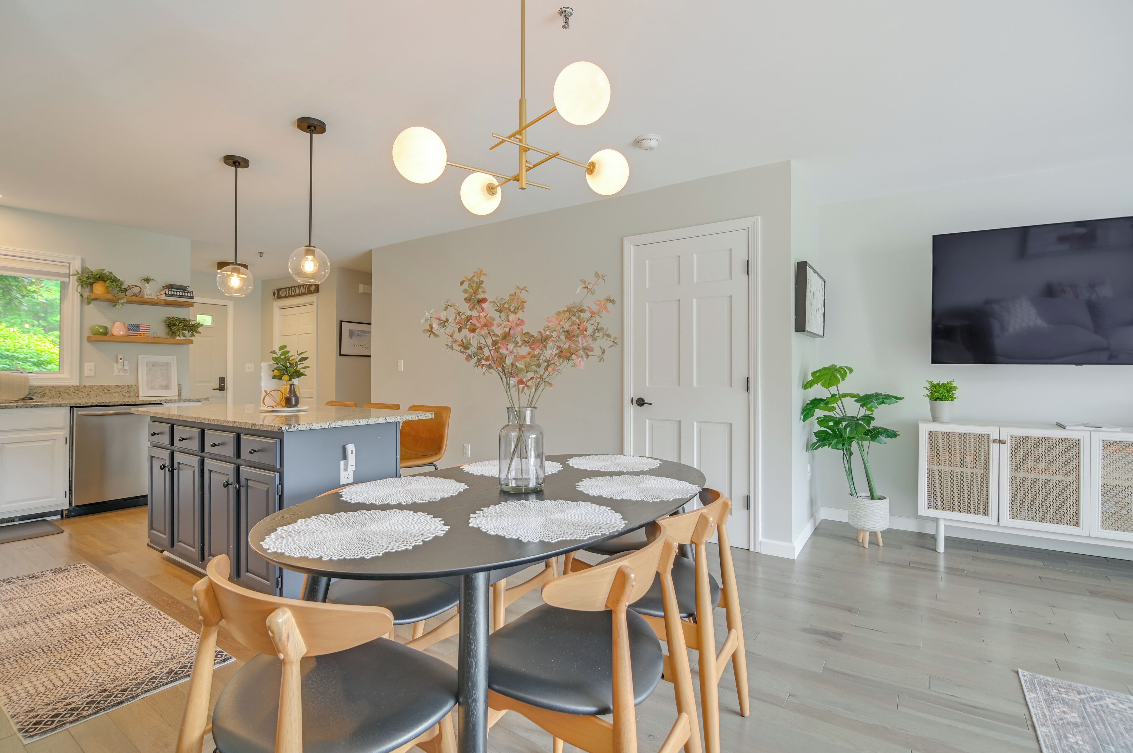 A modern kitchen with a dining area, wooden chairs, a round table with a white placemat, pendant lights, and a TV mounted on the wall.