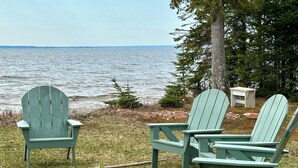 Two Adirondack chairs by a lakeside with a fire pit and a small table.