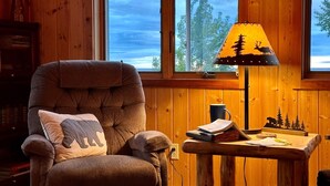 A cozy room with a large window, a comfortable armchair, a small wooden table with a lamp, and a bookshelf.