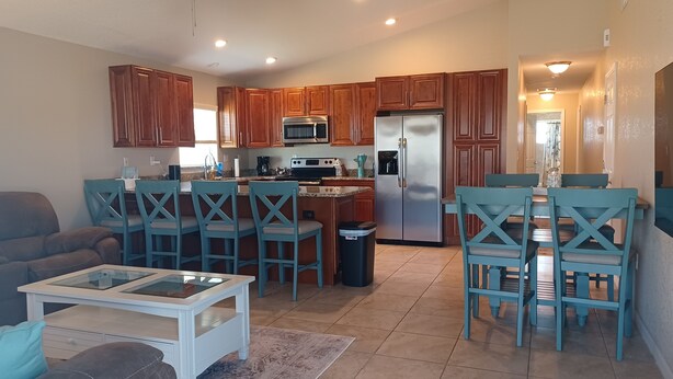A modern kitchen with wooden cabinets, a stainless steel refrigerator, and a dining area with blue chairs.