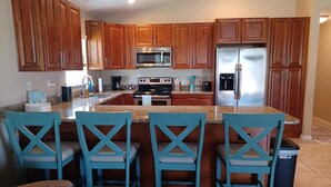 A kitchen with wooden cabinets, a stainless steel refrigerator, a microwave, and a coffee maker. There are four blue chairs around a central island.