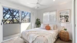 Un dormitorio con cama, ventilador de techo, ventana con persianas, una planta y un cuadro en la pared.