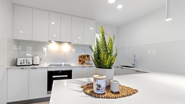 Una cocina moderna con muebles blancos, una isla central con helecho en maceta y un mantel tejido.