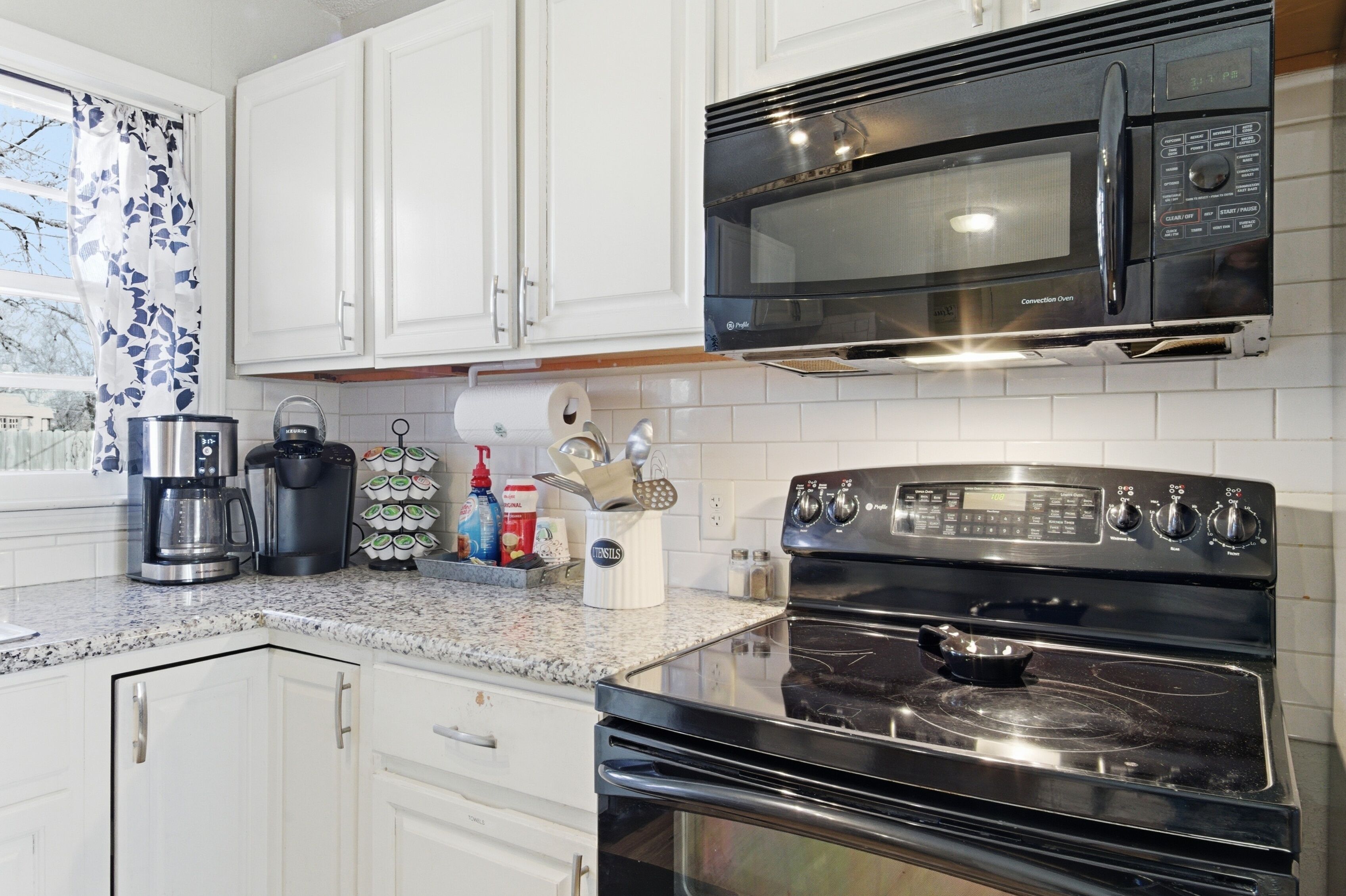A kitchen with white cabinets, a black microwave, a stove, and various kitchen items on the countertop.