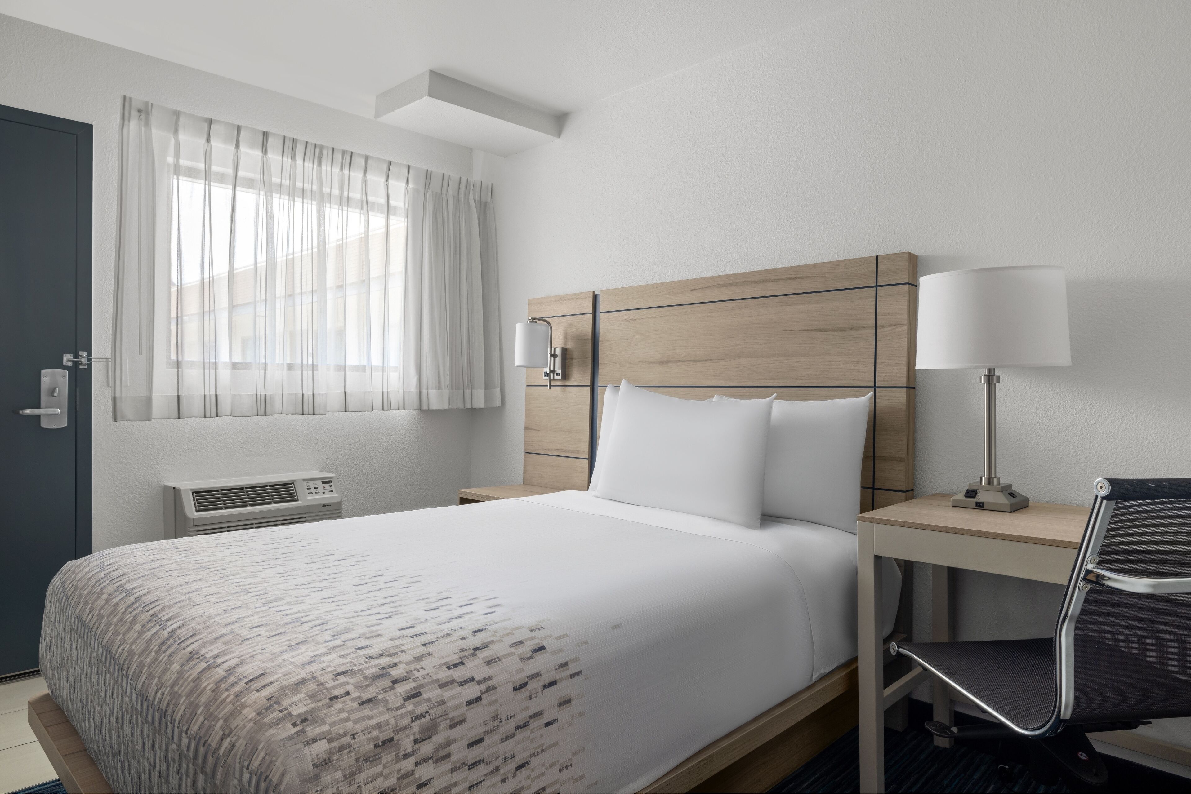 Una habitación de hotel con cama, escritorio, silla, lámpara, ventana con cortinas y un equipo de aire acondicionado de pared.