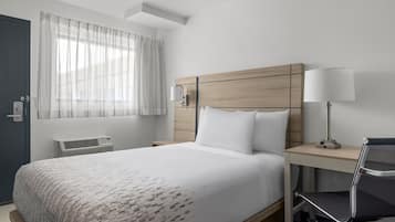 Una habitación de hotel con cama, escritorio, silla, lámpara, ventana con cortinas y un equipo de aire acondicionado de pared.
