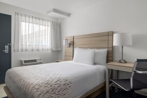 Una habitación de hotel con cama, escritorio, silla, lámpara, ventana con cortinas y un equipo de aire acondicionado de pared.