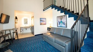 A hotel room with a blue carpet, a gray sofa, a dining area with a table and chairs, a kitchenette, a TV mounted on the wall, and a staircase with blue steps. - Orange Grove Inn (Tustin)