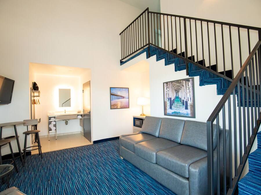 A hotel room with a blue carpet, a gray sofa, a dining area with a table and chairs, a kitchenette, a TV mounted on the wall, and a staircase with blue steps.
