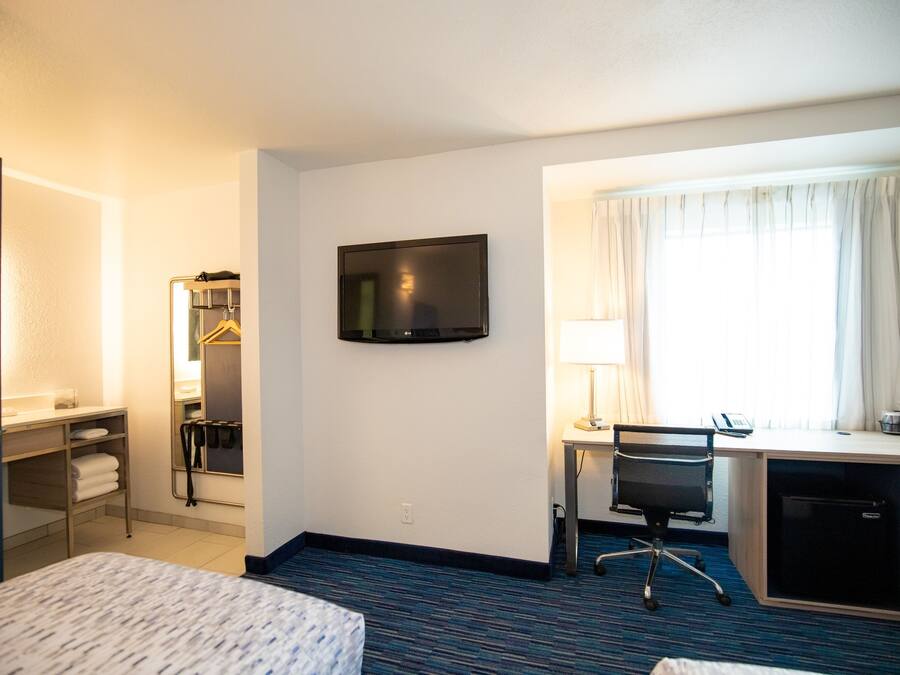 Habitación de hotel con cama, escritorio con computadora, televisor montado en la pared y ventana con cortinas.