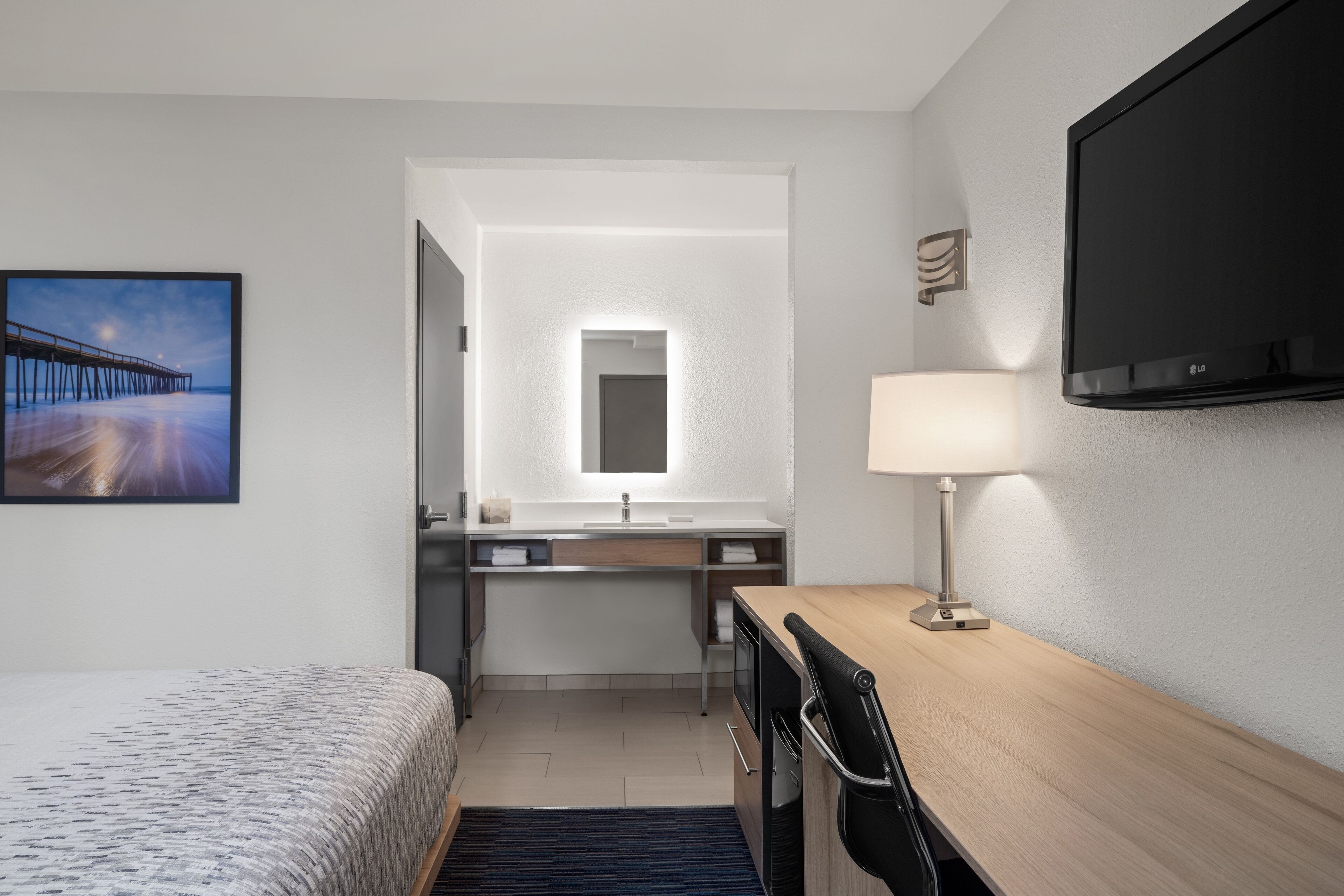 Una habitación de hotel con cama, escritorio con lámpara, televisión, baño con espejo y un cuadro de un muelle.