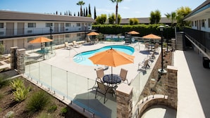 A pool area with lounge chairs and umbrellas, surrounded by a glass fence, and adjacent to a building with a balcony. - Orange Grove Inn (Tustin)