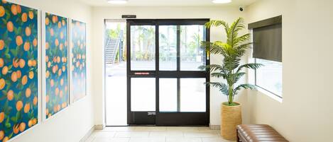 Un área de recepción con una puerta negra, un banco, una planta en maceta y una pared con cuadros de fruta naranja.