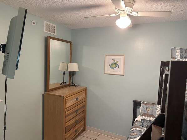 A bedroom with a wooden dresser, a ceiling fan, a framed picture on the wall, and a bed with patterned bedding.