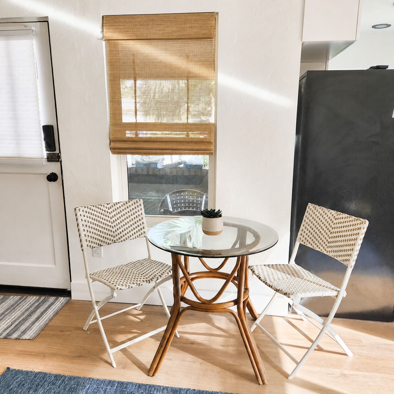 Un petit coin repas avec une table ronde en verre et deux chaises en osier, une fenêtre avec un store en bambou et un réfrigérateur noir.