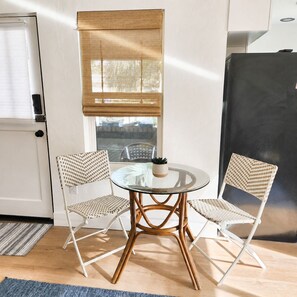 A small dining area with a round glass table and two wicker chairs, a window with a bamboo blind, and a black refrigerator. - Coastal Getaway 3-Min Walk to Beach & Sunsets (Treasure Island)