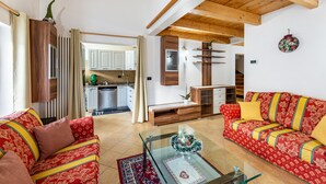 A living room with a sofa, a coffee table, a kitchen in the background, wooden beams on the ceiling, and a wall-mounted shelf.
