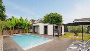 Ein moderner Poolbereich im Freien mit einem kleinen Gebäude, Liegestühlen und tropischen Pflanzen.